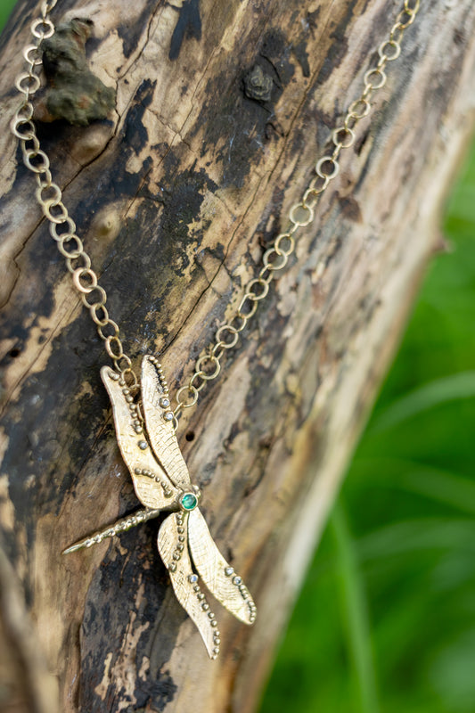 Yellow gold dragonfly pendant set with emerald and diamonds, hanging from handmade gold chain, with tree bark background