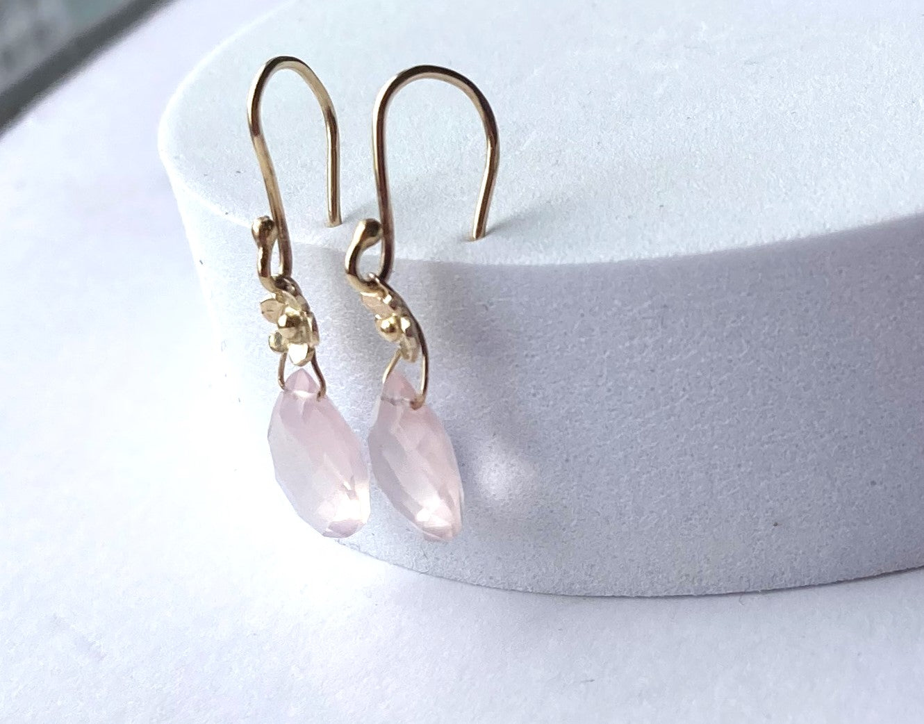 side view of gold floral drop earrings with pear shape rose quartz gemstones, stuck into top of white plinth