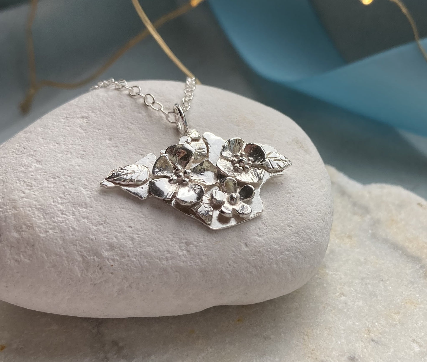 silver floral pendant silhouette of Isle of Wight, on white pebble with blue ribbon behind