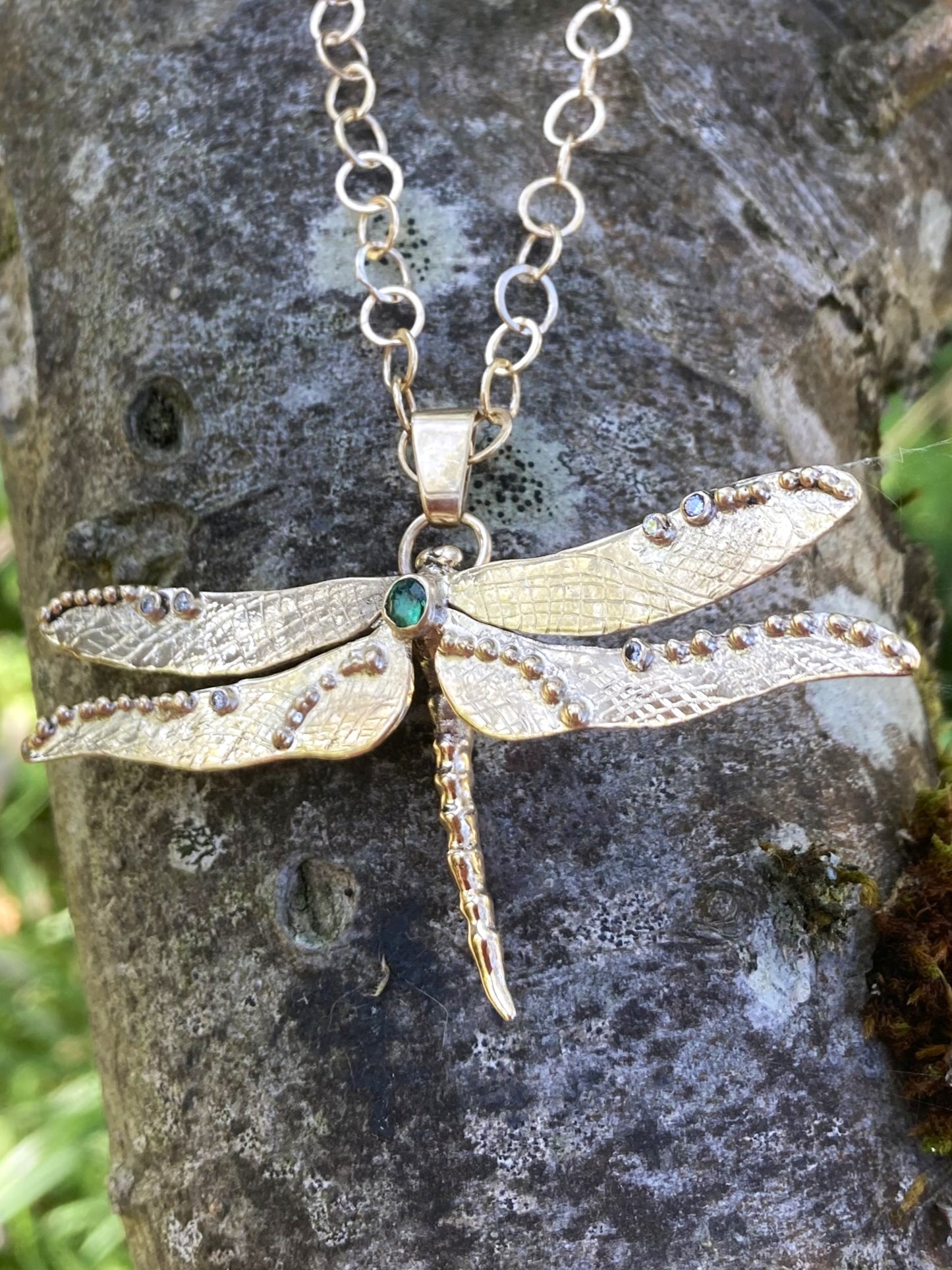 Gold dragonfly necklace with green emerald and diamonds, hanging on hand made gold chain, on tree background