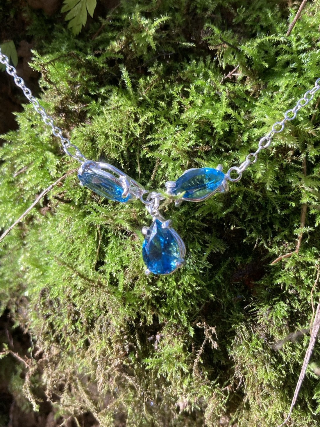 Blue teardrop-shaped necklace on a chain against a green moss background