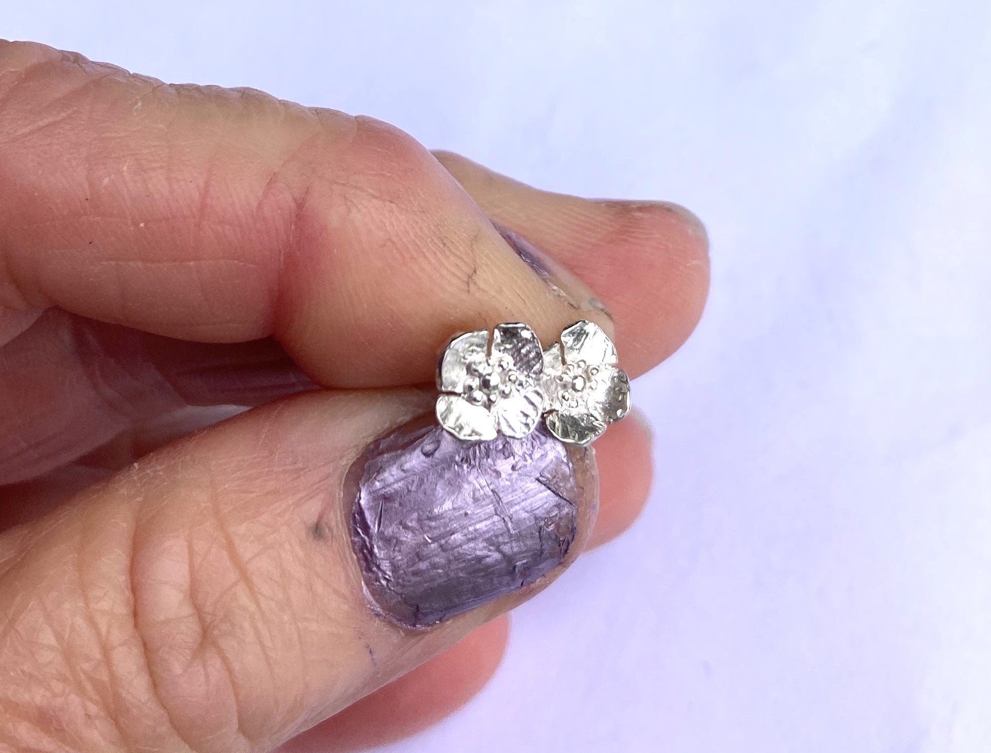 hand with purple nail varnish holding two small silver poppy stud earrings