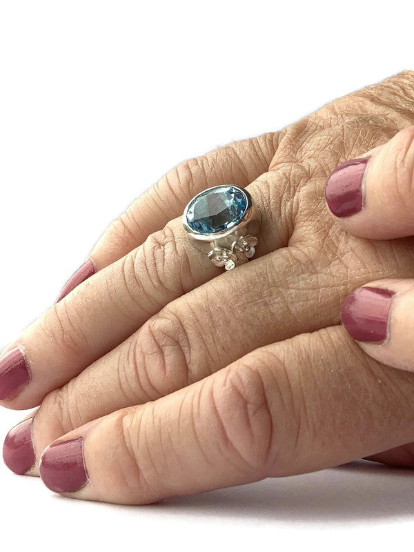 hand wearing a Kim Styles floral cocktail ring set with a large oval blue topaz with floral details, on a white background