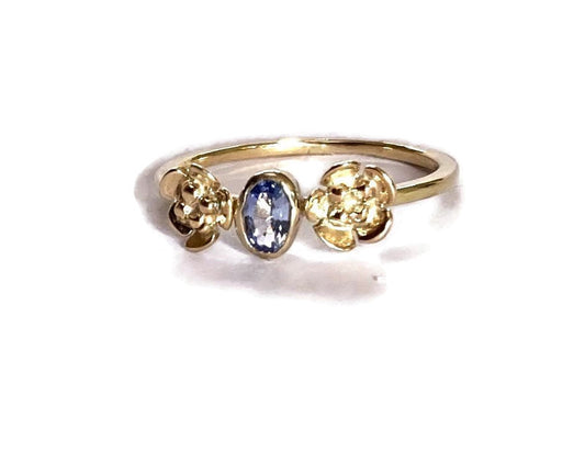 Pretty gold ring with flowers either side of central gemstone and set with light blue oval sapphire, alternative engagement ring,  on white background