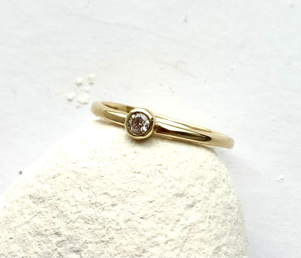 Kim Styles 18ct yellow gold round diamond solitaire ring with halo band, leaning on white pebble