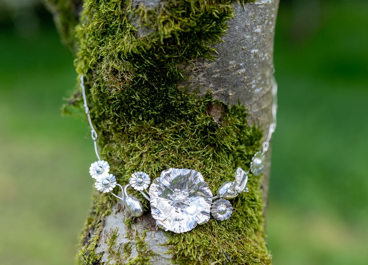 large silver necklace with hand forged textured poppy and daisy theme, on mossy tree