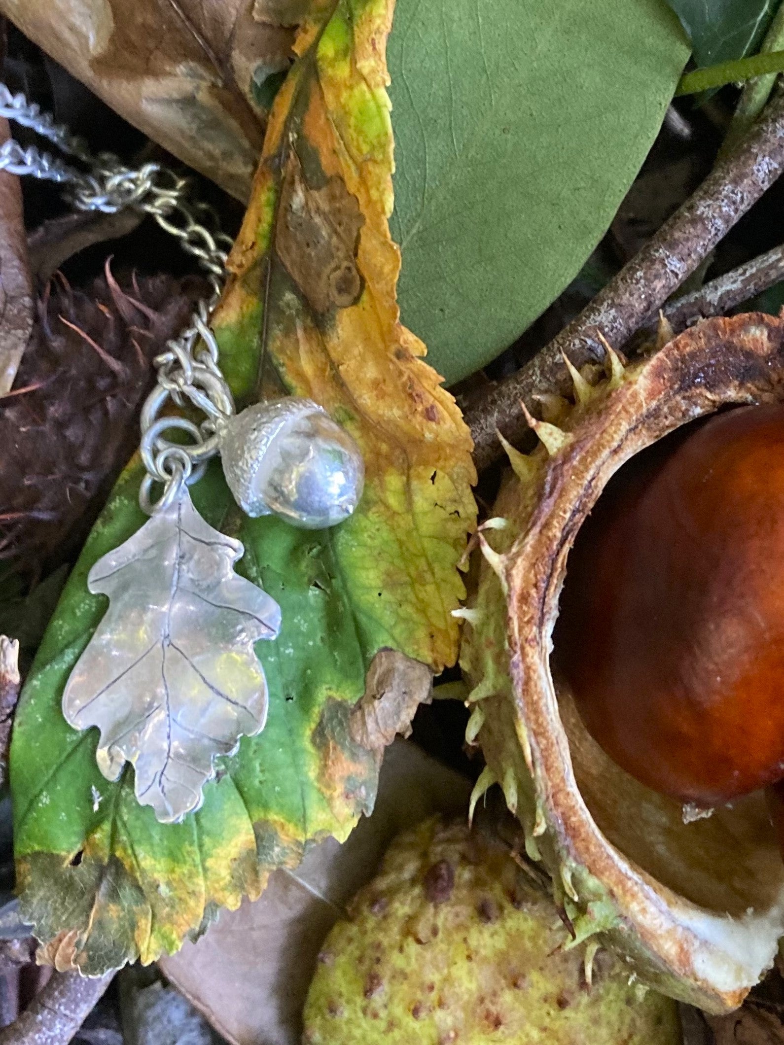 Tiny silver acorn and hand forged oak leaf pendant in sterling silver, resting on autumn leaves and a conker by Kim Styles Jewellery on the Isle of Wight
