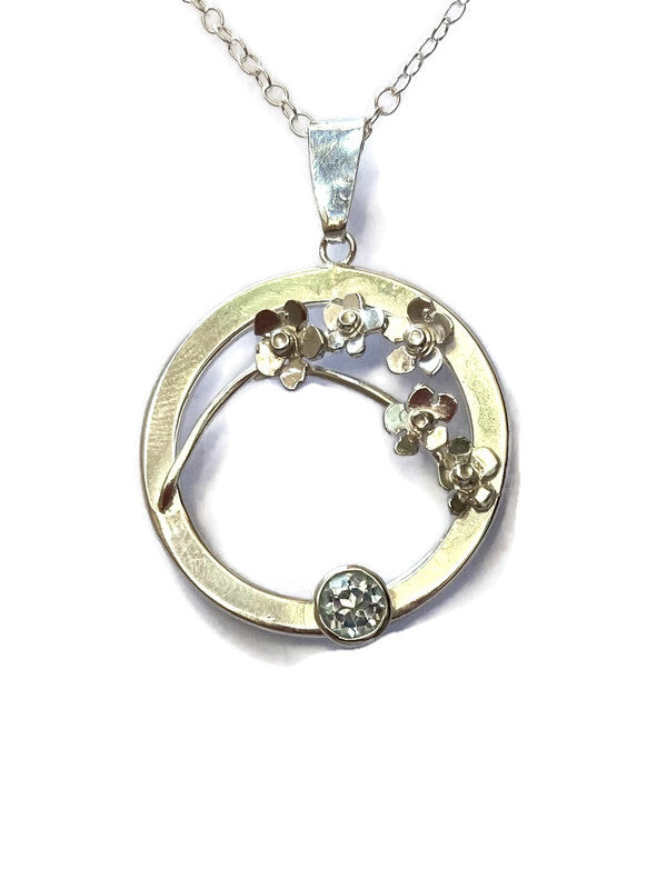 circle silver pendant with sprig of tiny silver forget-me-not flowers and round pale blue  gemstone on white background