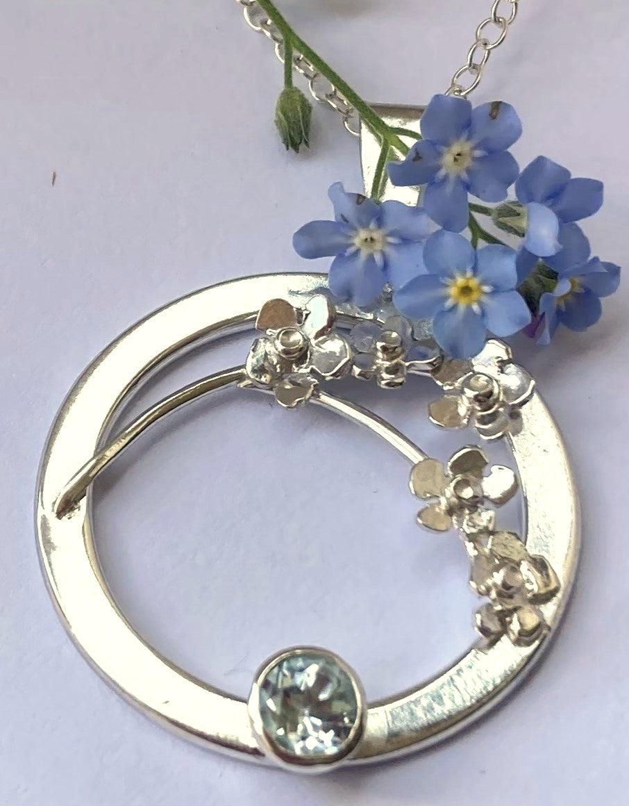 sprig of real blue forget-me-not flowers resting on silver circle pendant with silver forget-me-not flowers and set with round blue topaz
