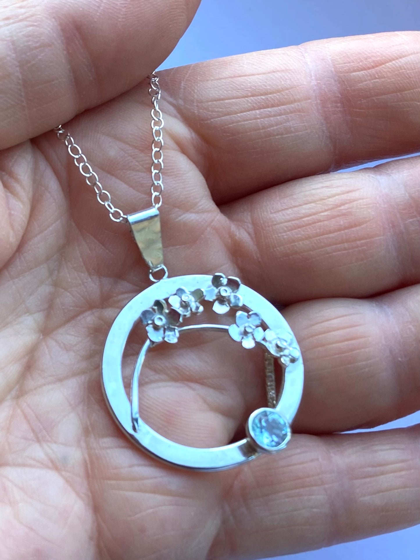 hand holding silver circle and flowers pendant set with pale blue gemstone