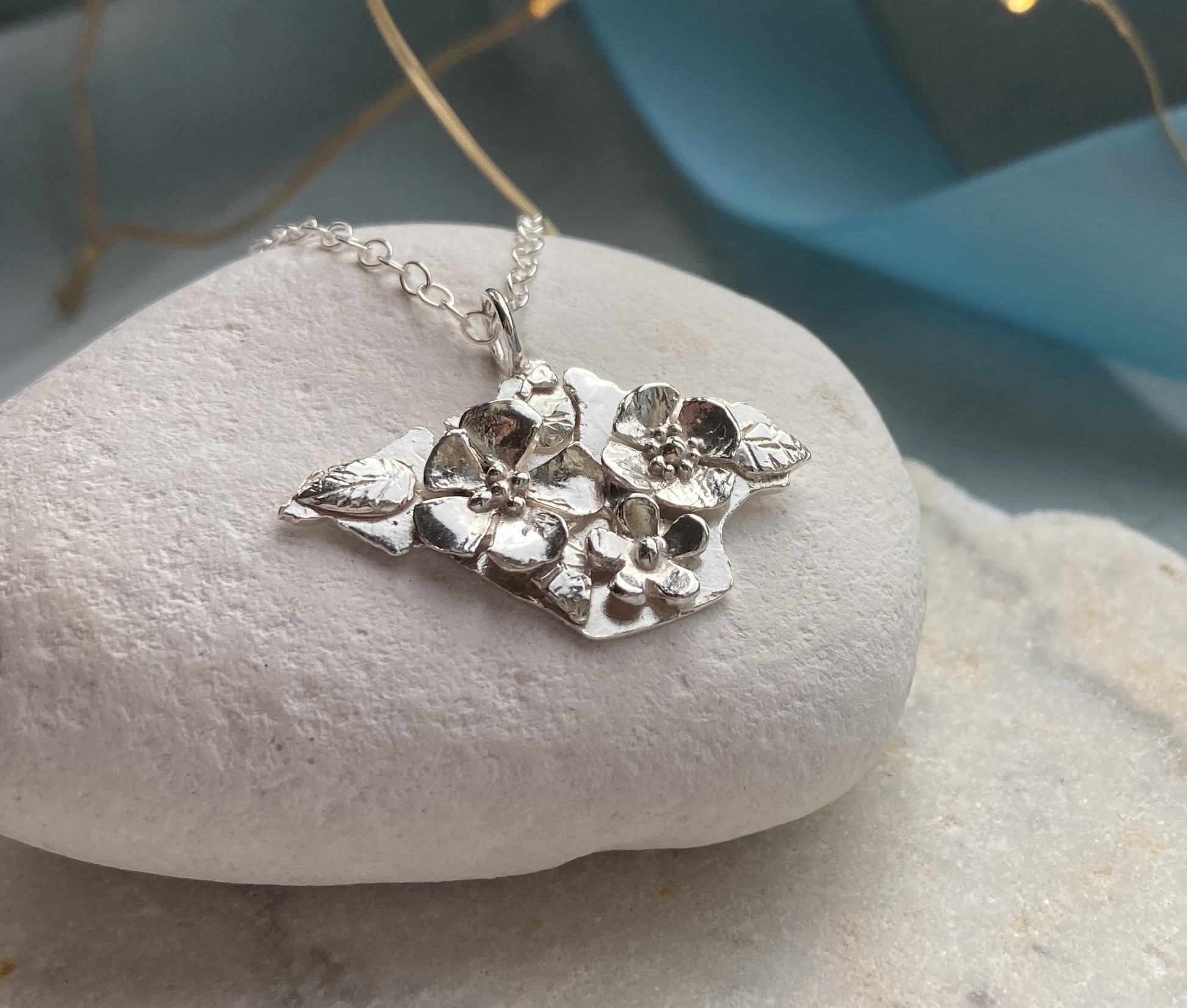 silver floral pendant silhouette of Isle of Wight, on white pebble with blue ribbon behind
