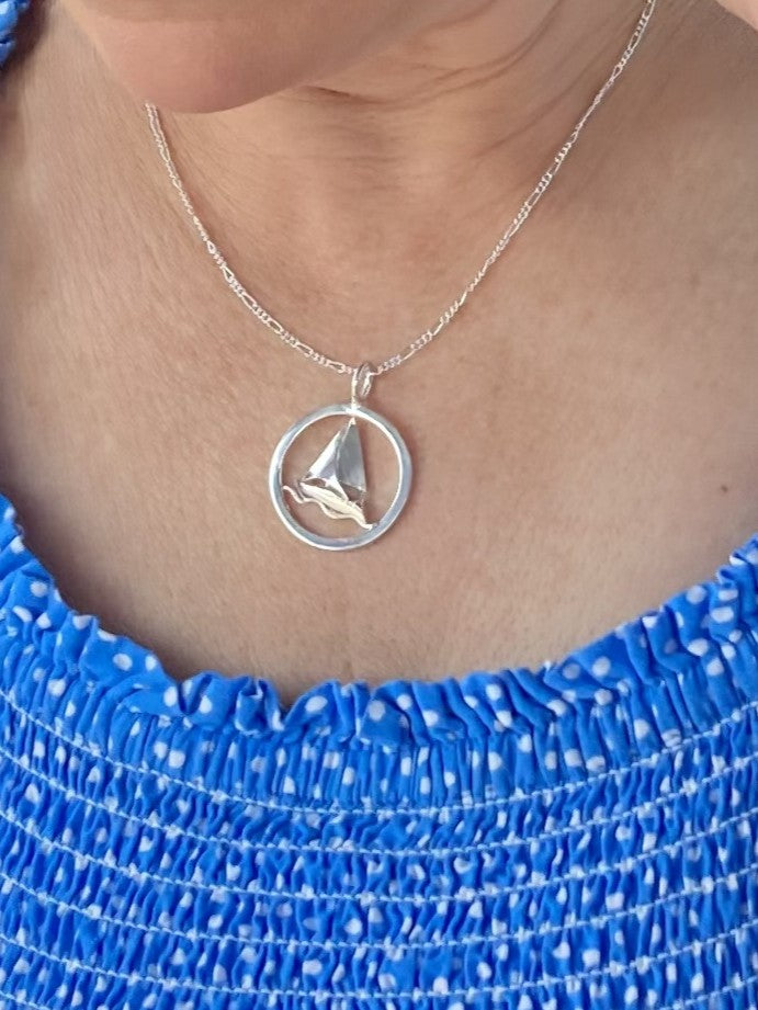 A close-up of a person wearing a delicate silver chain necklace with a circular pendant featuring a small  3D sailboat motif, paired with a blue and white patterned top.
