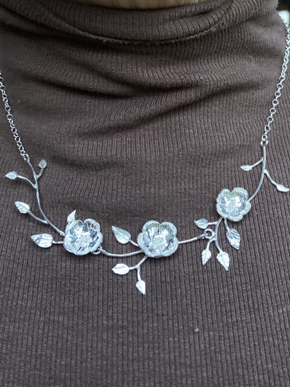A hand forged organic sterling silver necklace with three textured flower motifs and delicate leafy vines draped across a dark brown ribbed turtleneck sweater.