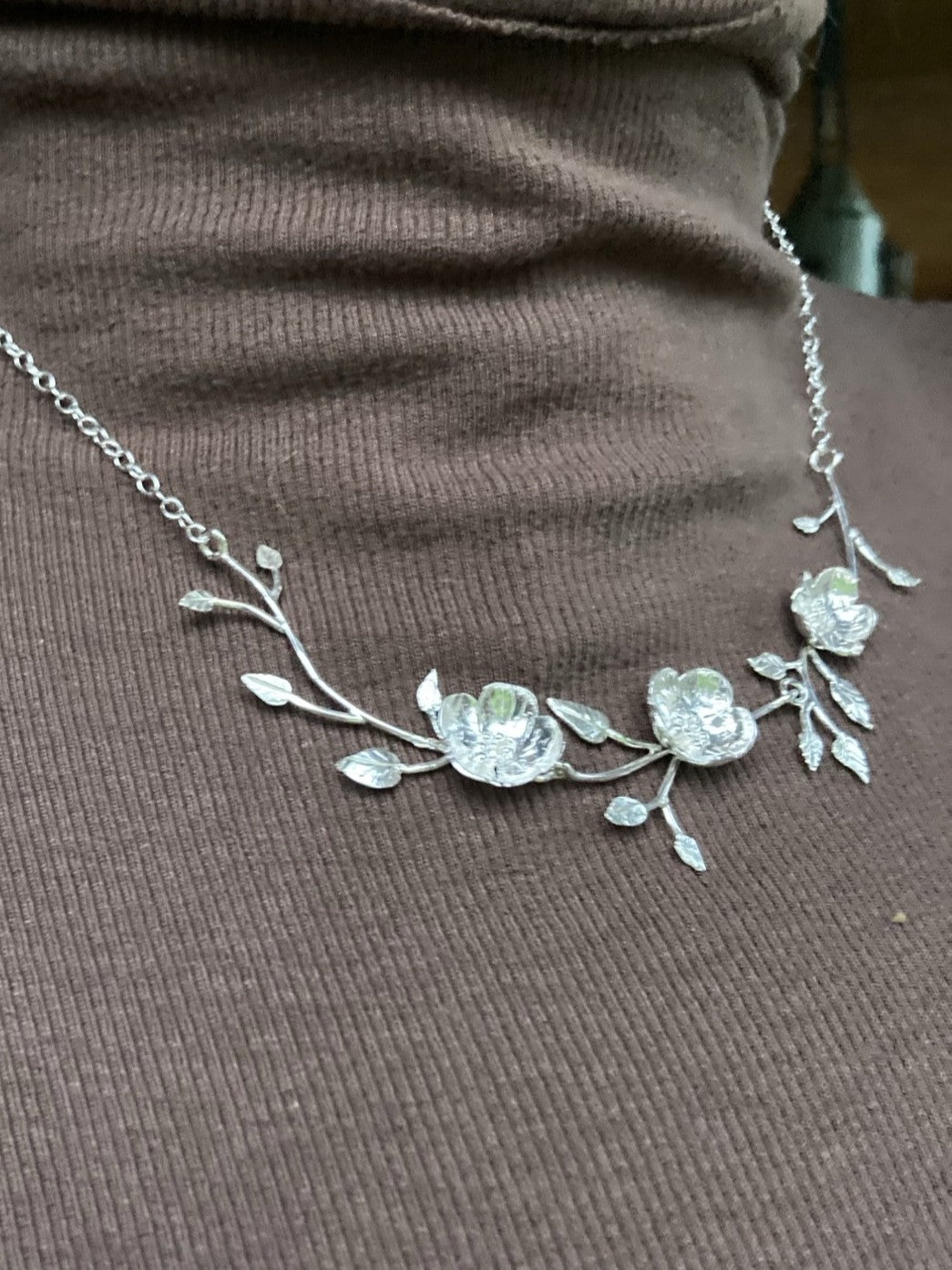 A sterling silver floral branch necklace with three textured flower motifs and leaves, displayed on a ribbed brown turtleneck sweater.