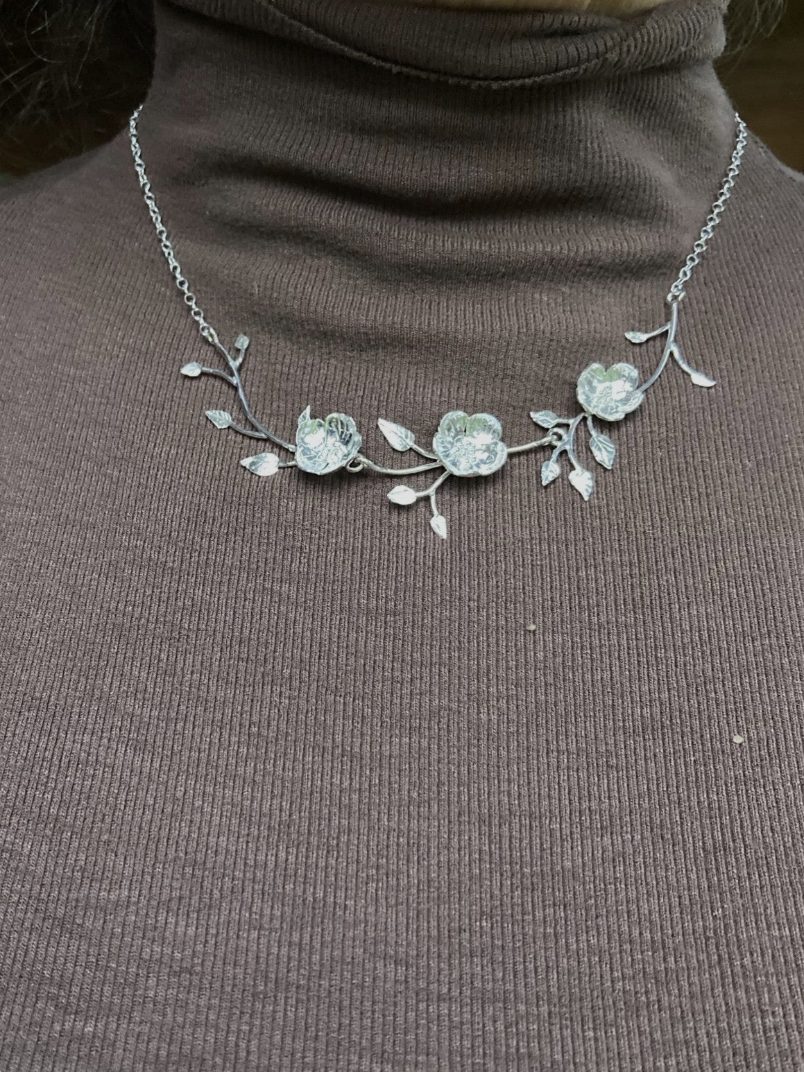 A silver floral branch necklace with three wild rose flowers, displayed on a person's chest over a brown ribbed turtleneck sweater.