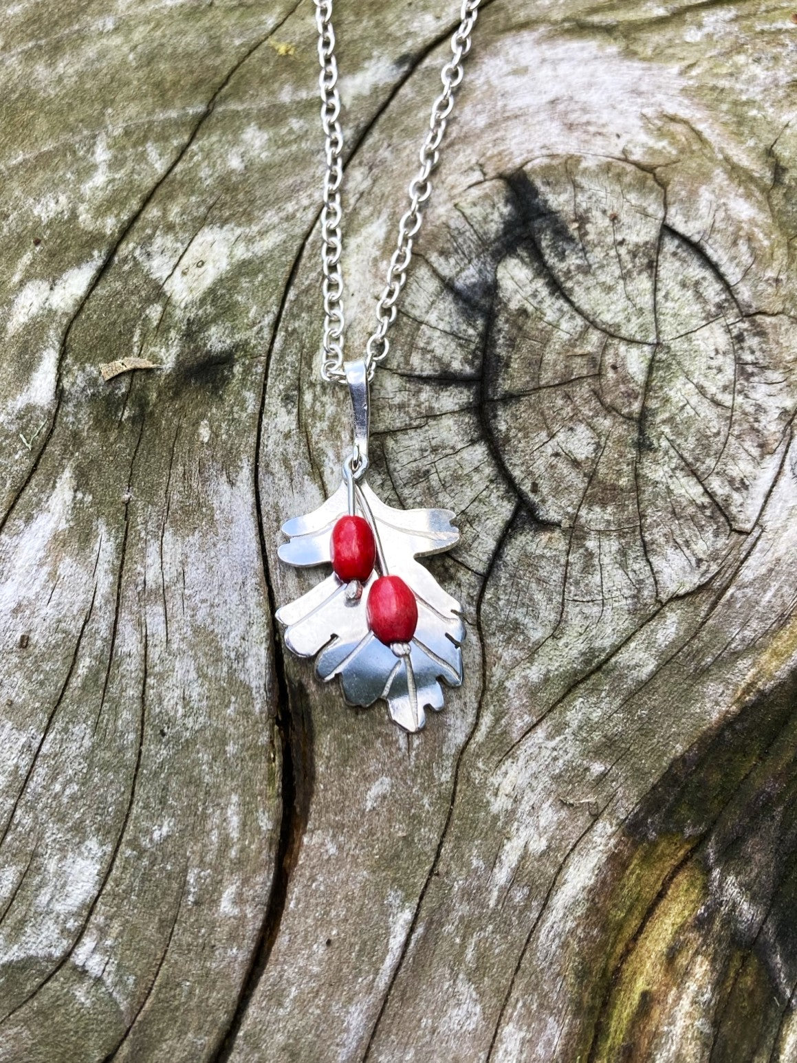 A silver  Hawthorne leaf pendant necklace with two red berry beads, displayed on a weathered wood surface.