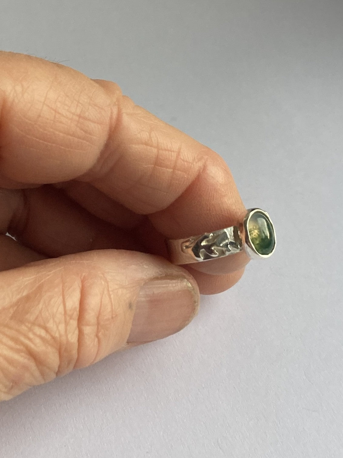 A close-up of a hand holding a leaf detail silver ring with a moss agate green gemstone against a plain light grey background.