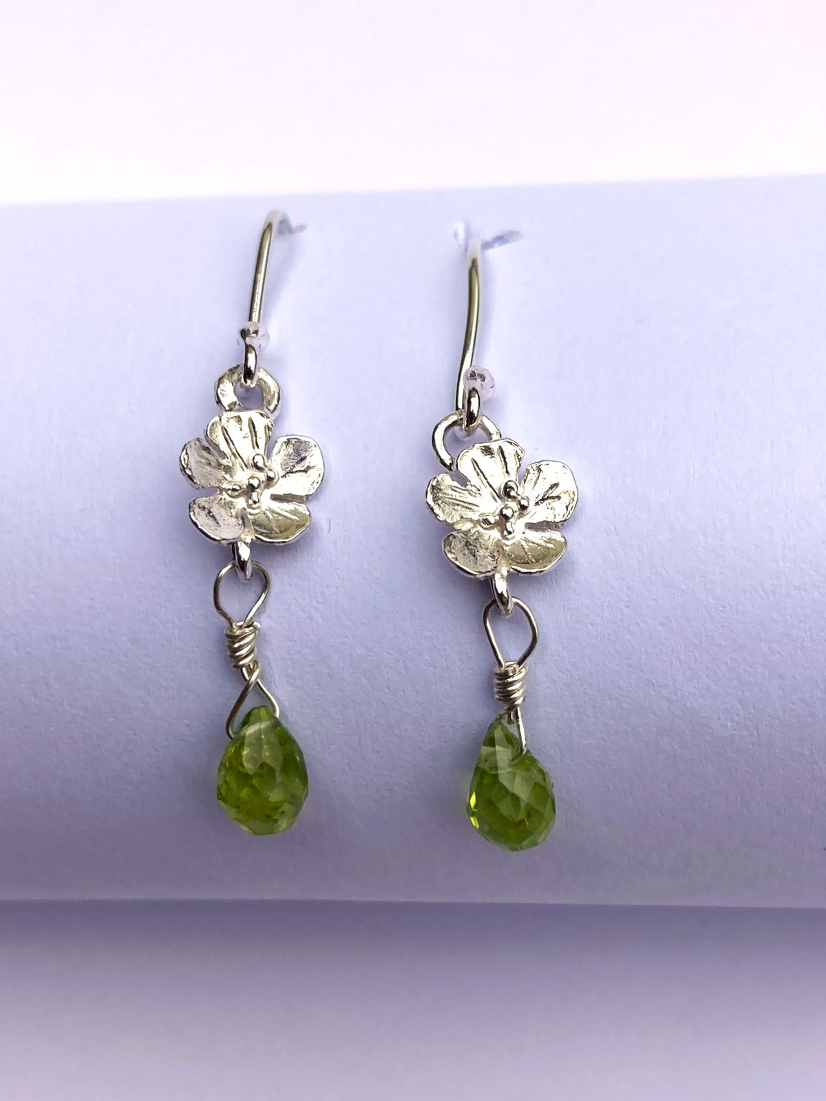 A pair of silver dangle earrings with textured flower charms and faceted green peridot 
teardrop gemstones, displayed on a light purple cylindrical stand.