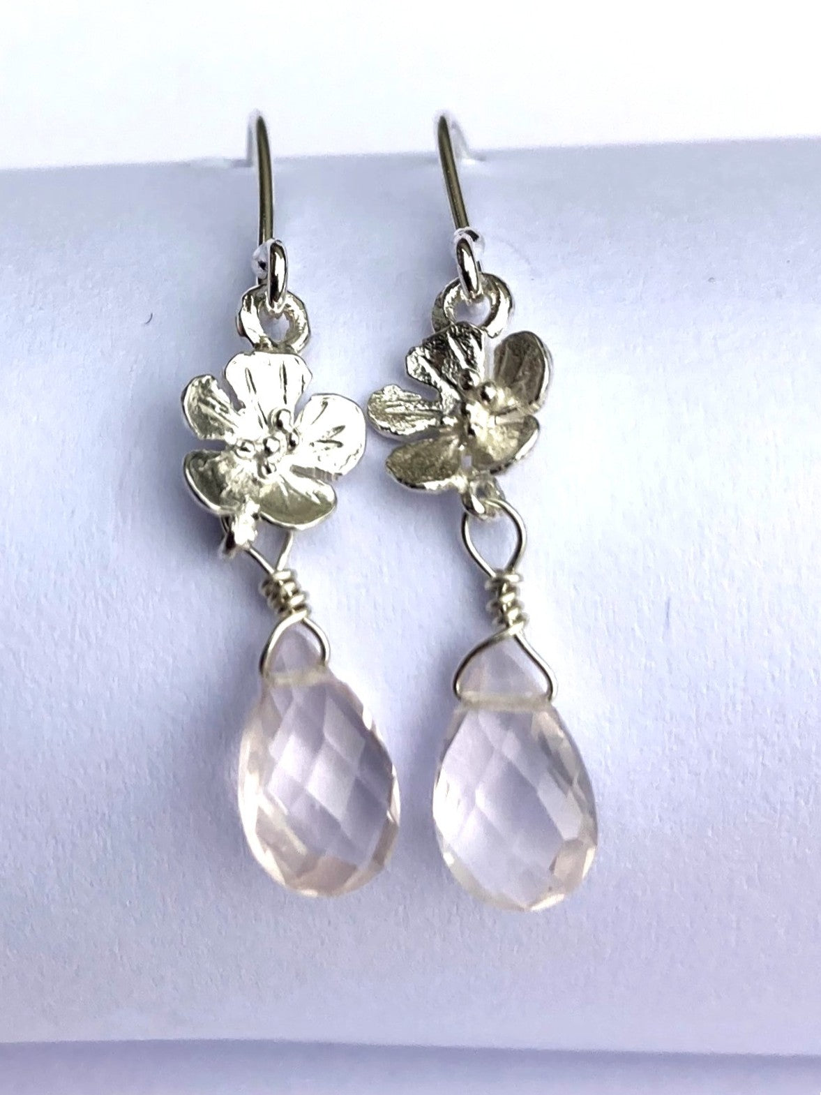 A pair of sterling silver dangle earrings with textured flower charms and faceted pink teardrop rose quartz drops, displayed on a white cylindrical stand.