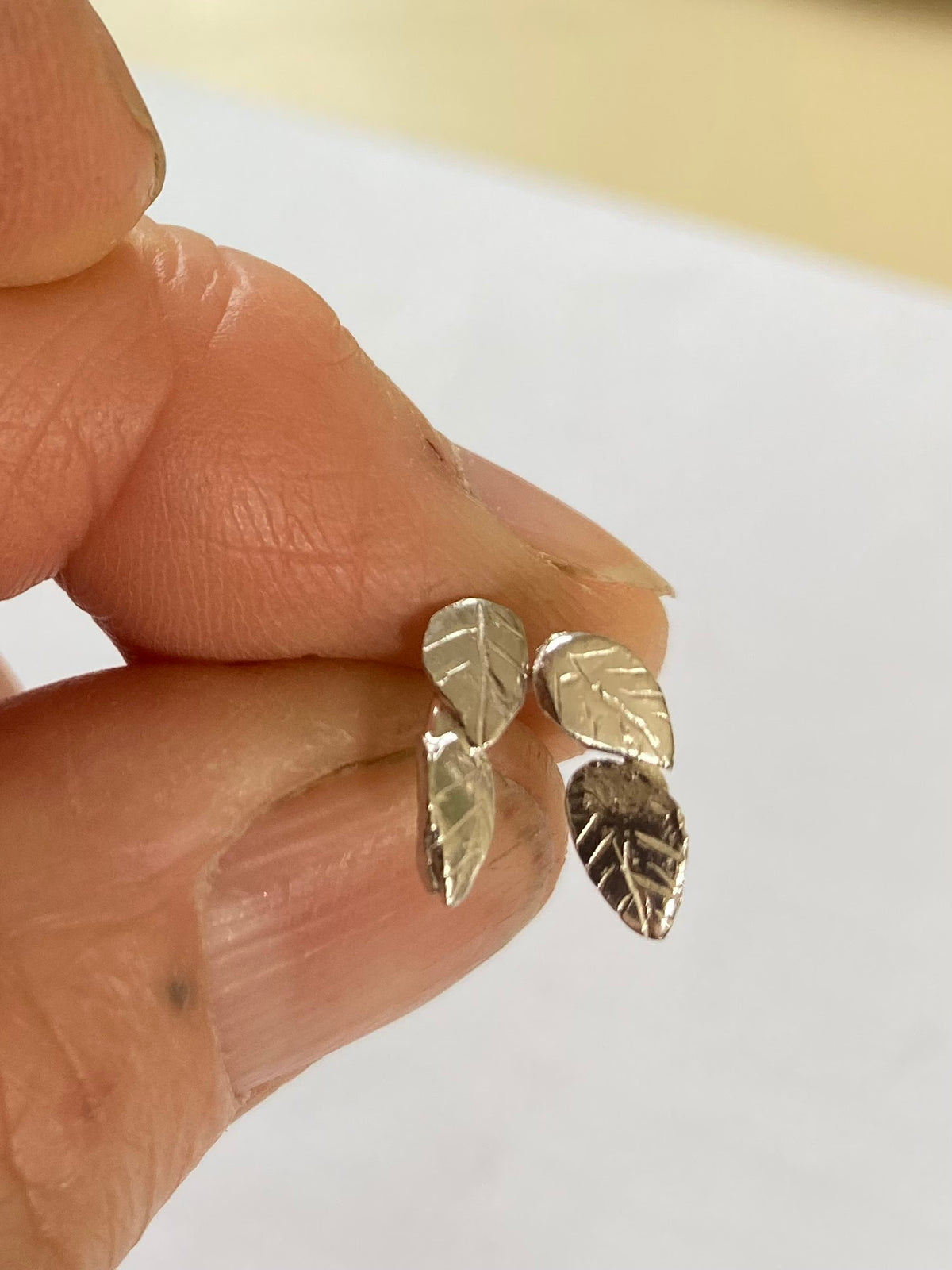 A close-up of a hand holding a pair of small silver leaf stud earrings against a plain light background.