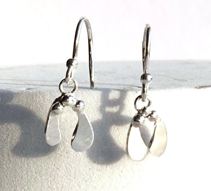 A pair of silver drop mistletoe leaf earrings, displayed on a textured white surface with soft shadows.