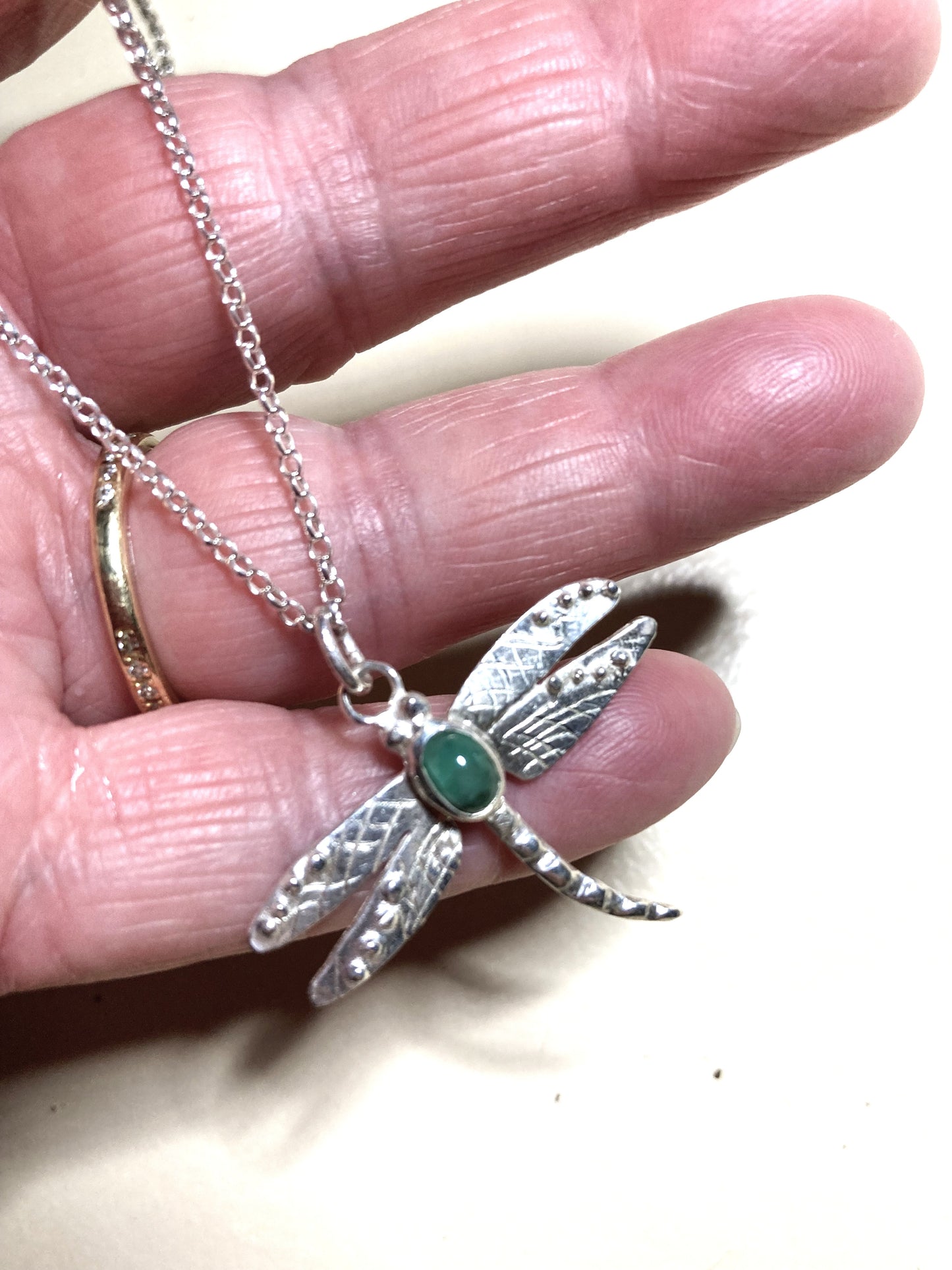 Silver dragonfly pendant with green oval gemstone held in a hand against a neutral background