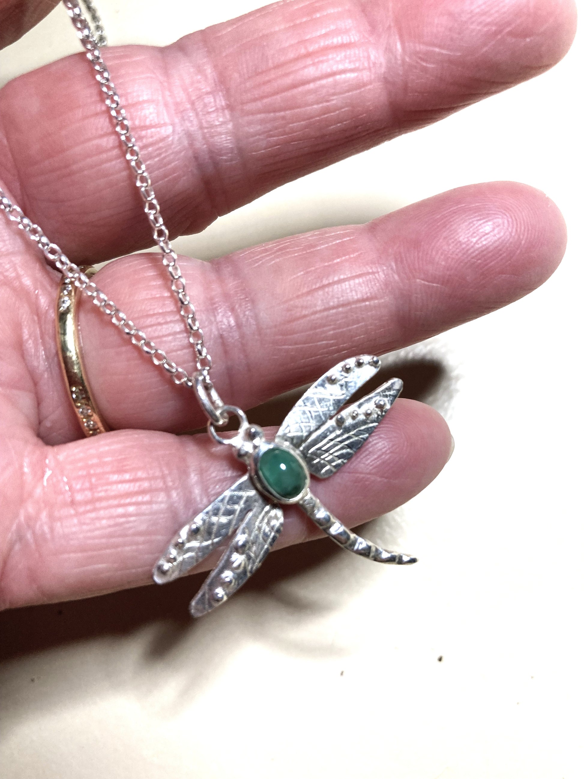 Silver dragonfly pendant with green oval gemstone held in a hand against a neutral background