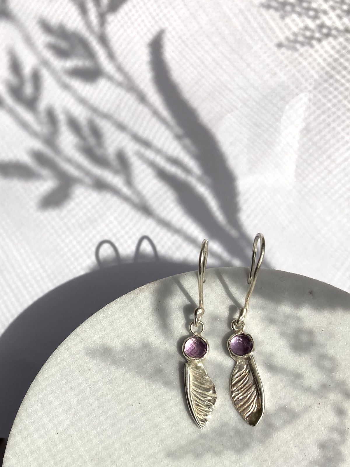handmade textured  silver sycamore seed hook earrings by Kim Styles Jewellery set with round chequerboard cut amethysts, on patterned white background