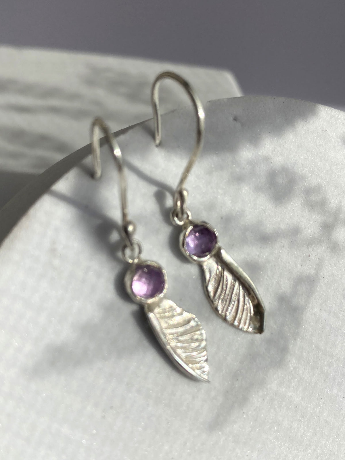 hand forged textured silver sycamore seed hook earrings by Kim Styles Jewellery set with round chequerboard cut amethysts, on patterned grey background