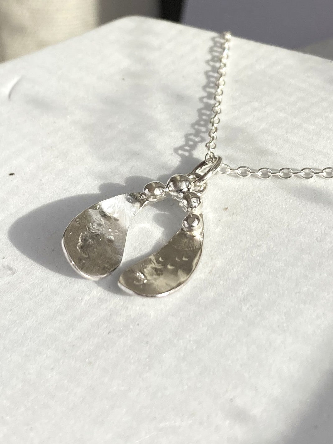 A silver mistletoe necklace with two textured, organic-shaped leaves on a delicate chain, displayed on a white textured surface.