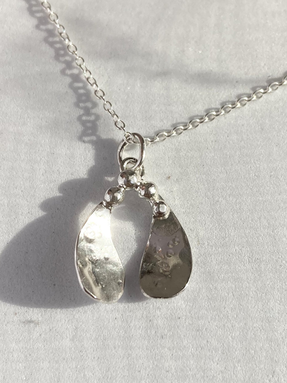 A silver mistletoe pendant necklace with a textured, hammered finish and a small cluster of silver berries at the top, shown on a light grey textured background with a soft shadow.