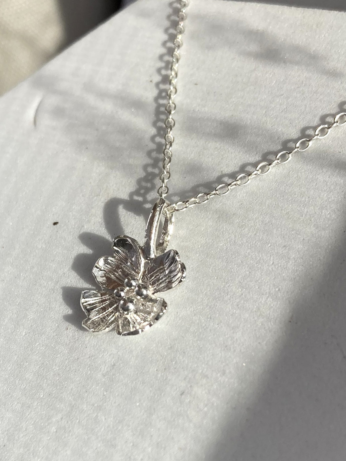 A silver  dogwood blossom flower pendant necklace on a delicate silver chain, resting on a white textured surface with soft shadows.