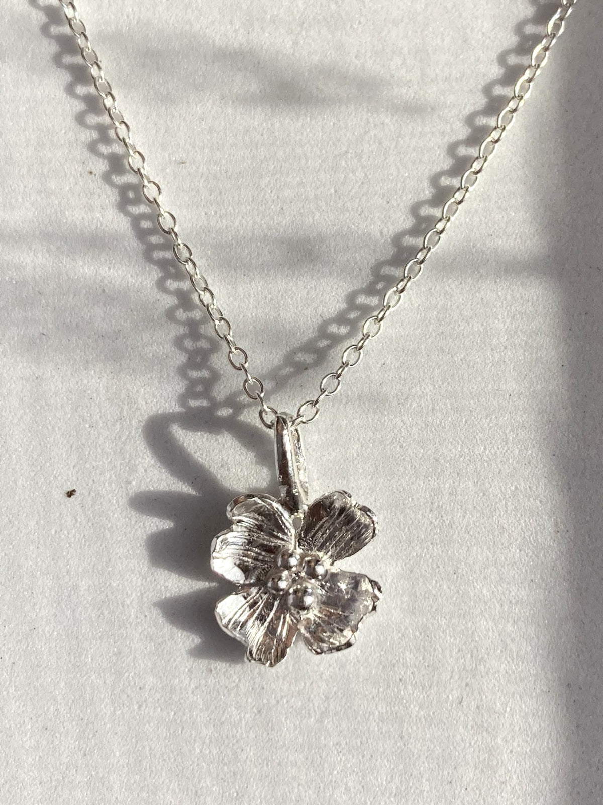 A hand forged silver Dogwood flower pendant necklace on a delicate silver chain, by Kim Styles Jewellery,  displayed on a textured white surface with soft leaf shadows.