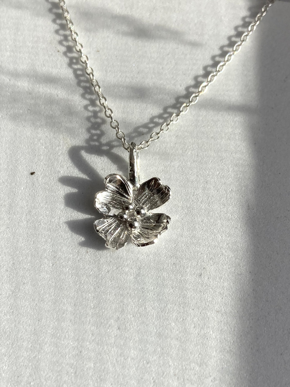 A silver Dogwood flower pendant necklace on a delicate silver chain, by Kim Styles Jewellery,  displayed on a textured white surface with soft leaf shadows.