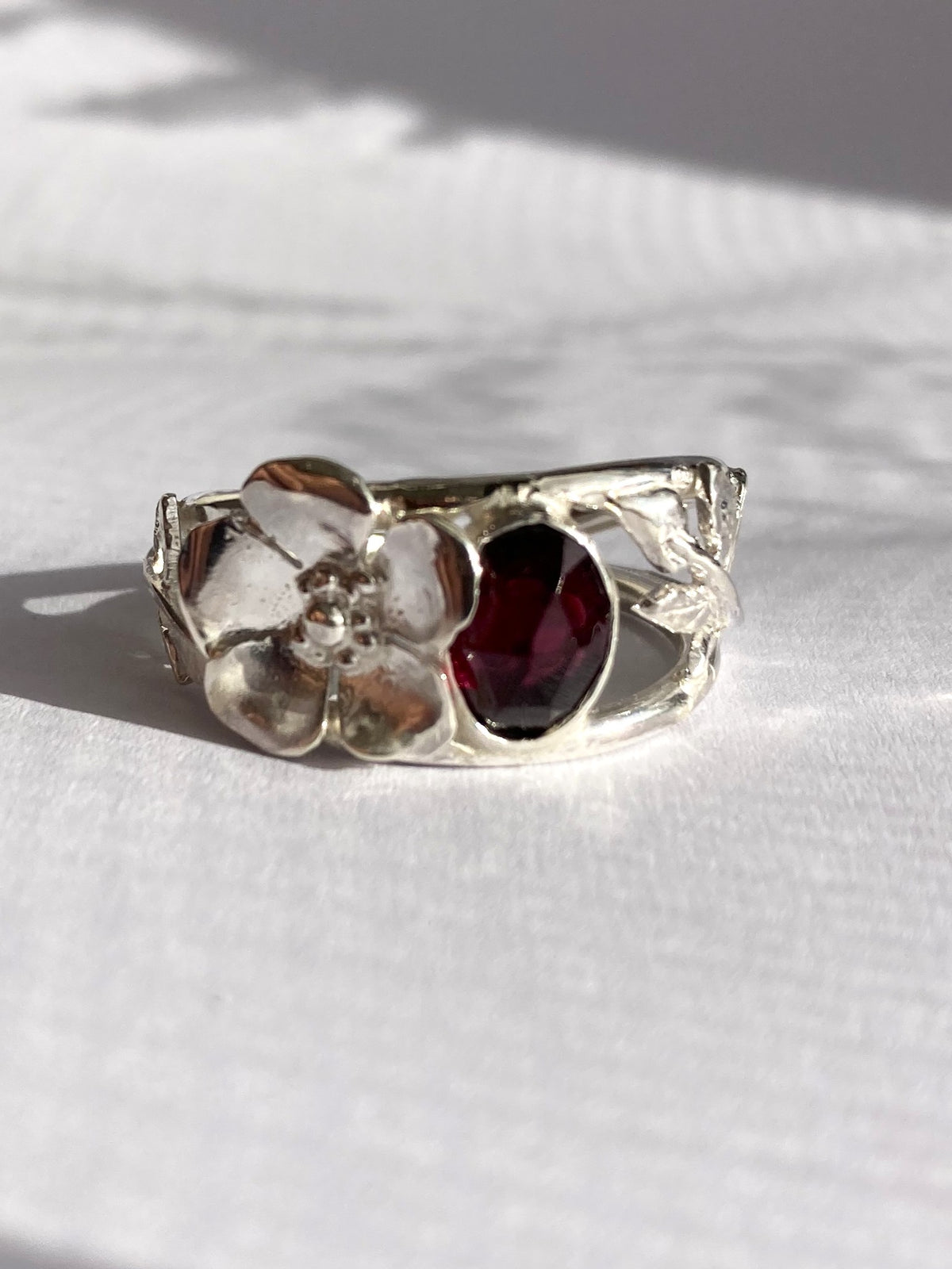 A silver ring featuring a sculpted flower with a deep red oval garnet gemstone, resting on a textured white surface with soft shadows.