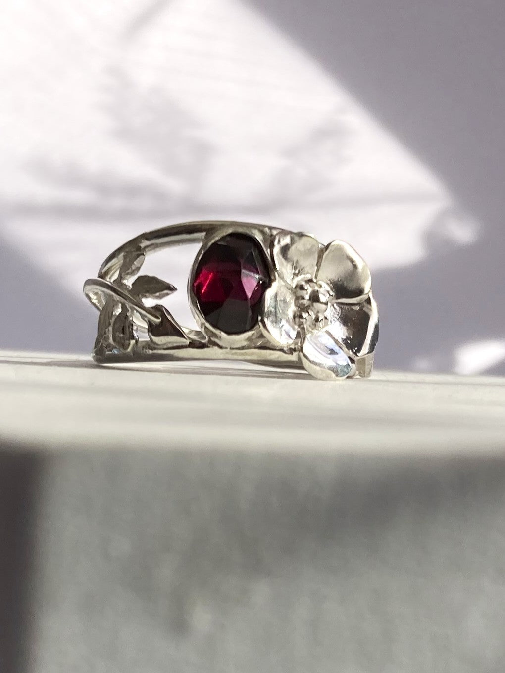 A silver ring by Kim Styles Jewellery featuring a central oval dark red gemstone and a floral design with a clear faceted stone, displayed on a light grey surface.