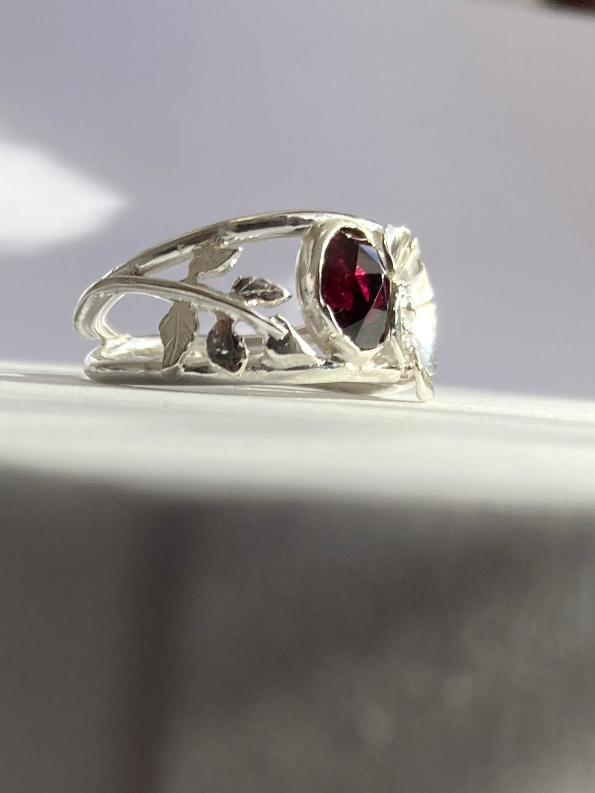 A silver ring with a floral design and a central rose cut oval red gemstone, resting on a light grey surface.