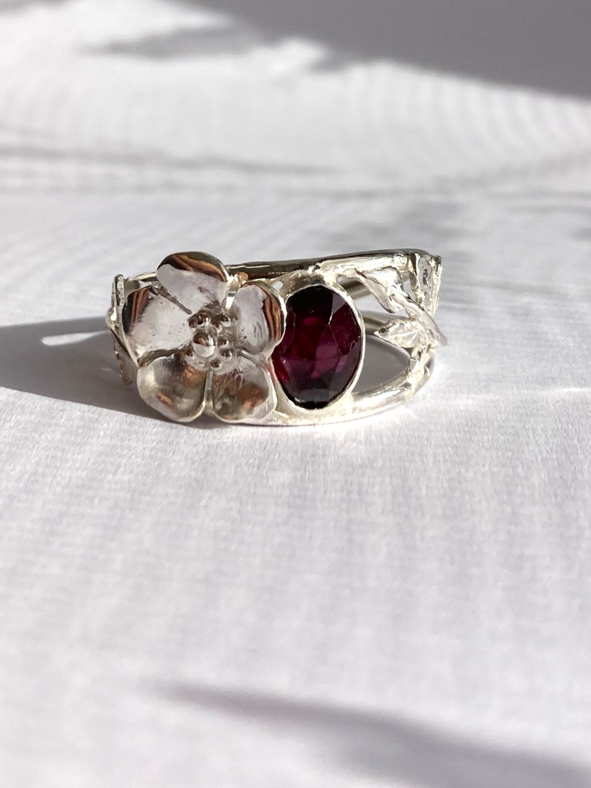 A silver ring featuring a sculpted flower and a central oval dark red garnet gemstone, resting on a textured white fabric surface.