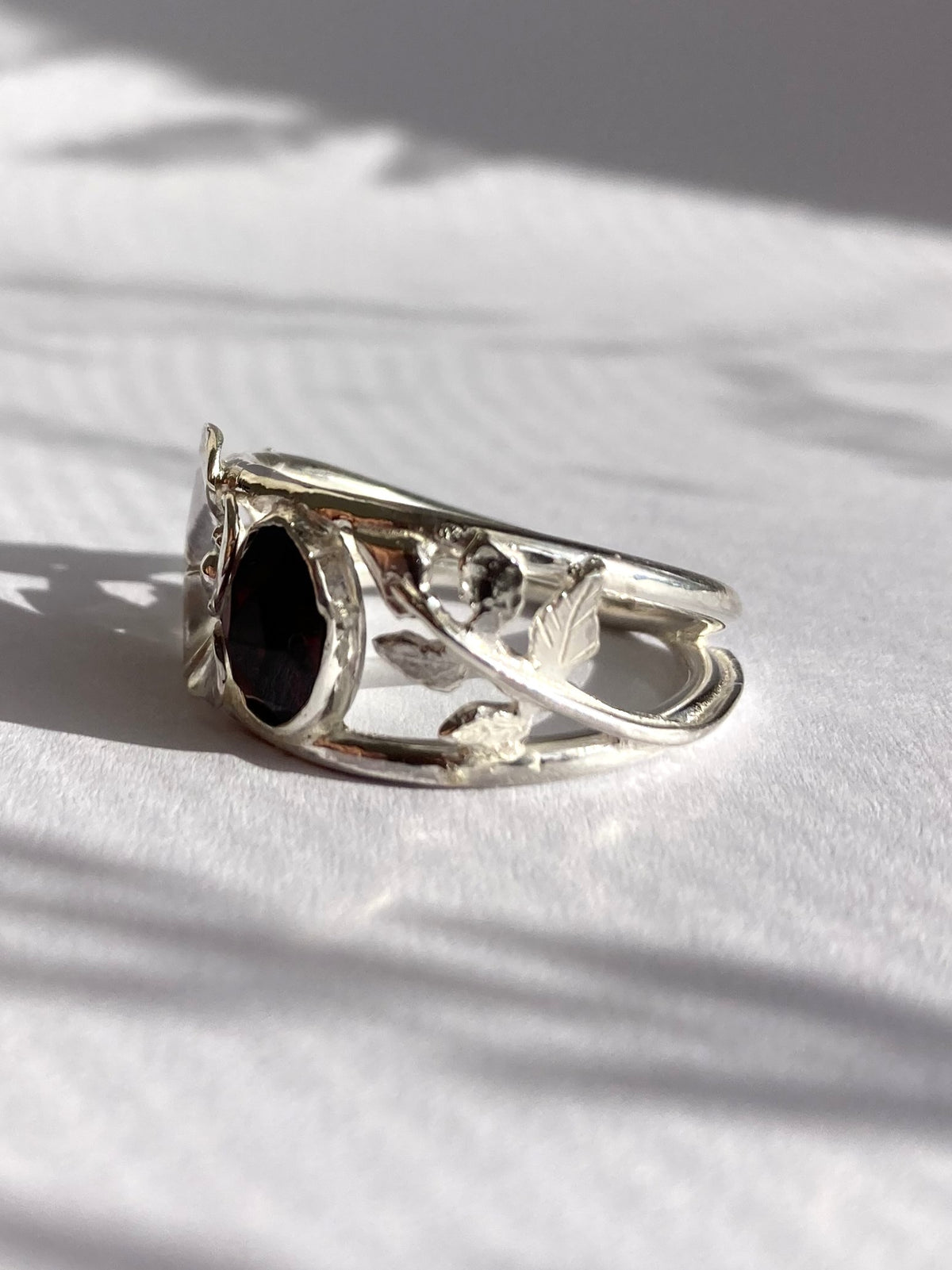 A silver ring with a dark oval garnet stone and delicate leaf-like detailing, resting on a white textured surface with soft shadows.
