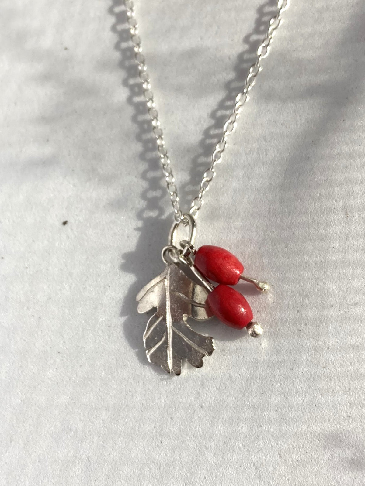 A silver necklace with a leaf and berry pendant, featuring two small red bead accents, displayed on a textured light grey fabric background.