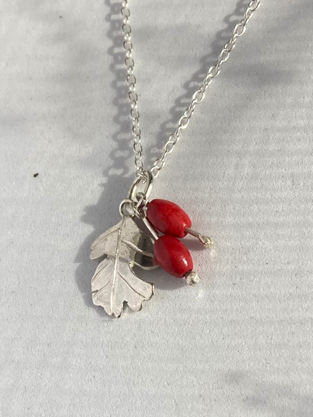 A silver chain necklace featuring a leaf charm and two small red berry-like beads, photographed on a light gray fabric background.