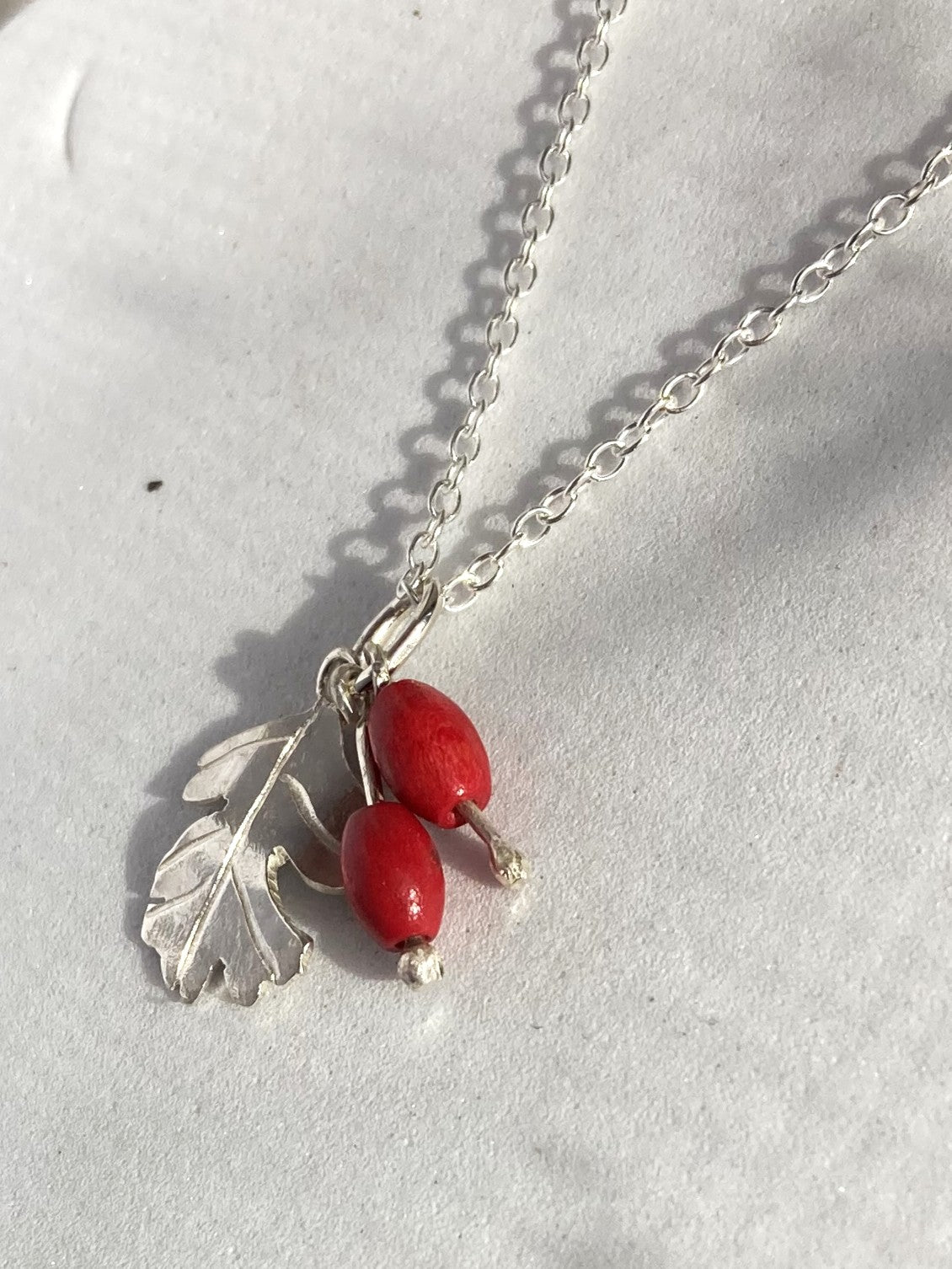 A silver necklace by Kim Styles Jewellery featuring a Hawthorne leaf and berry pendant with two small red beads, displayed on a textured white surface.