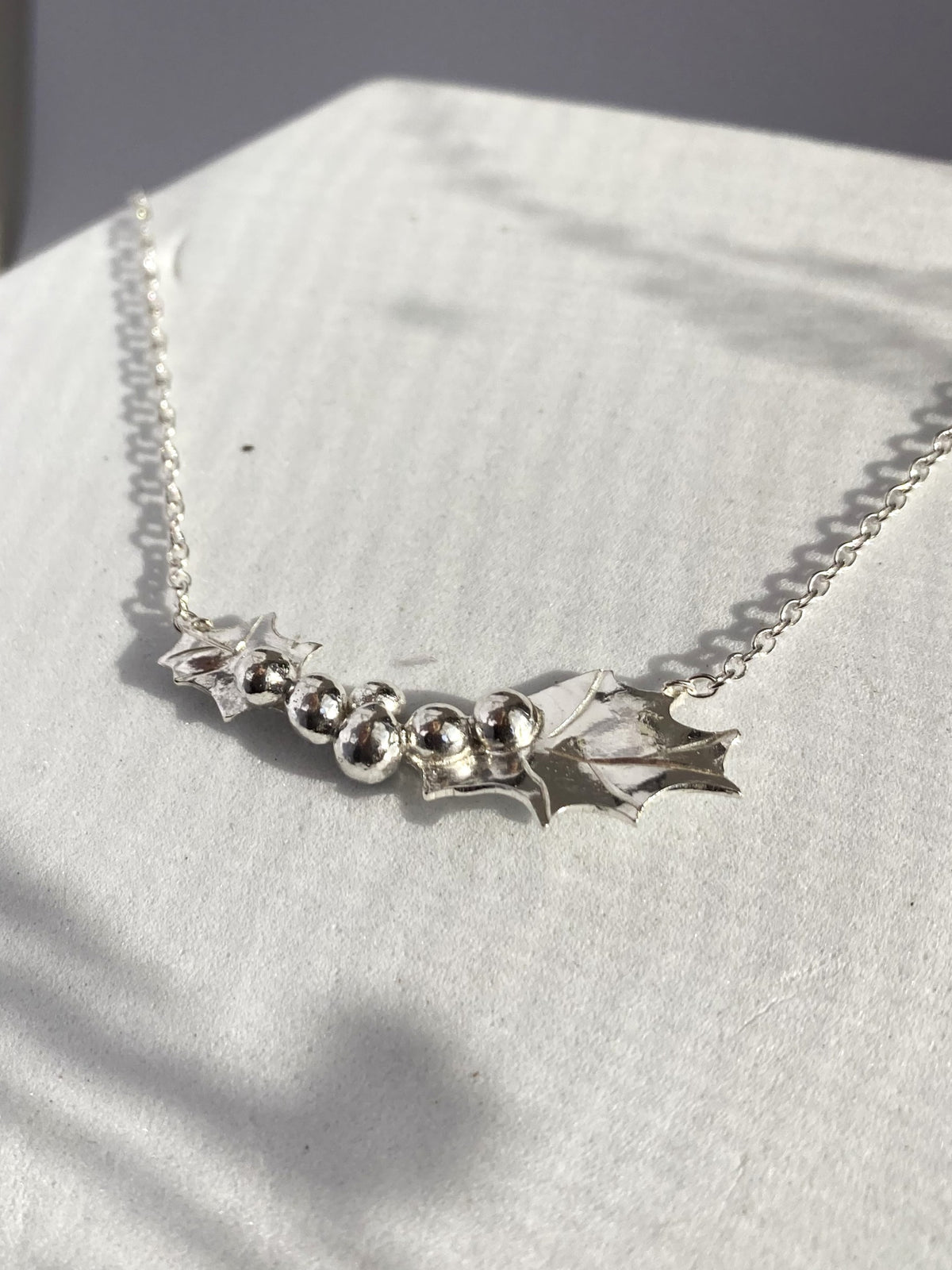 a sterling silver handmade holly leaf necklace with central clustered berries, on a grey textured background
