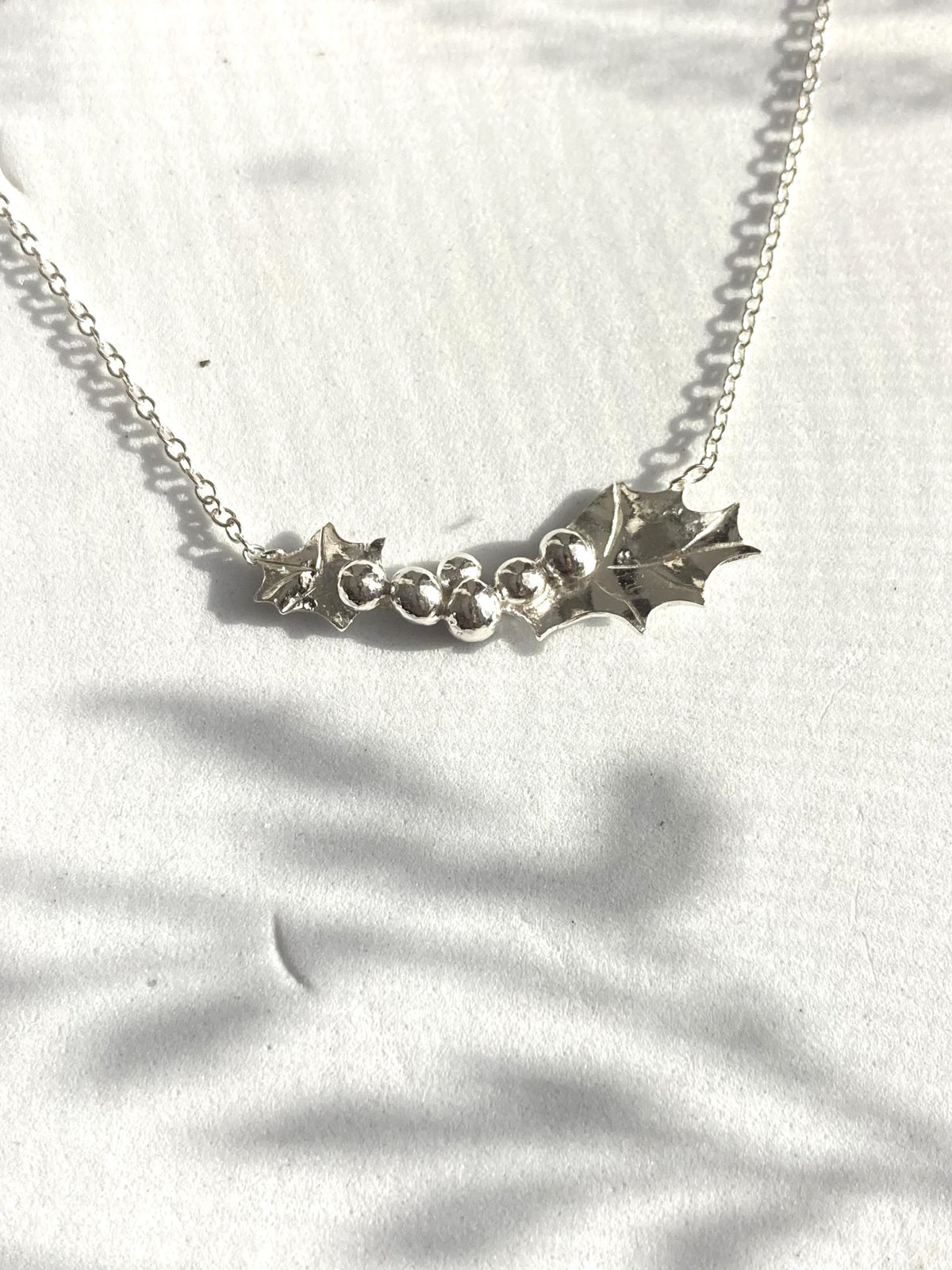 A silver Holly leaf necklace with a delicate chain and a central cluster of silver berries, displayed on a white textured fabric with soft shadow overlays.