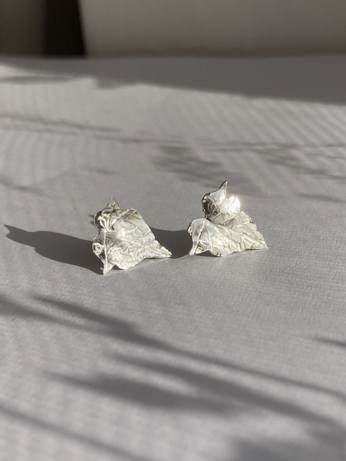 A pair of textured silver earrings shaped like ivy leaves, resting on a light grey fabric surface with soft shadows cast across it.
