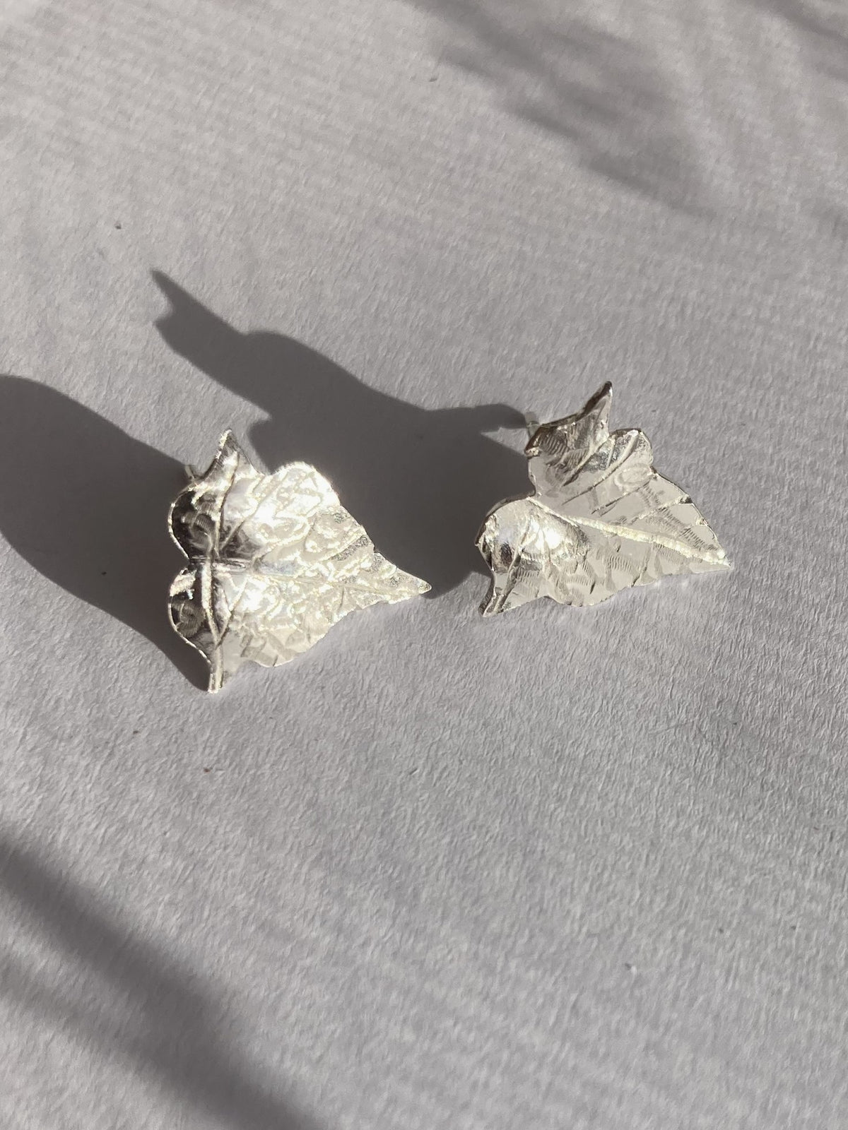 A pair of textured silver ivy leaf-shaped stud earrings resting on a light grey fabric surface with soft shadows cast across it.