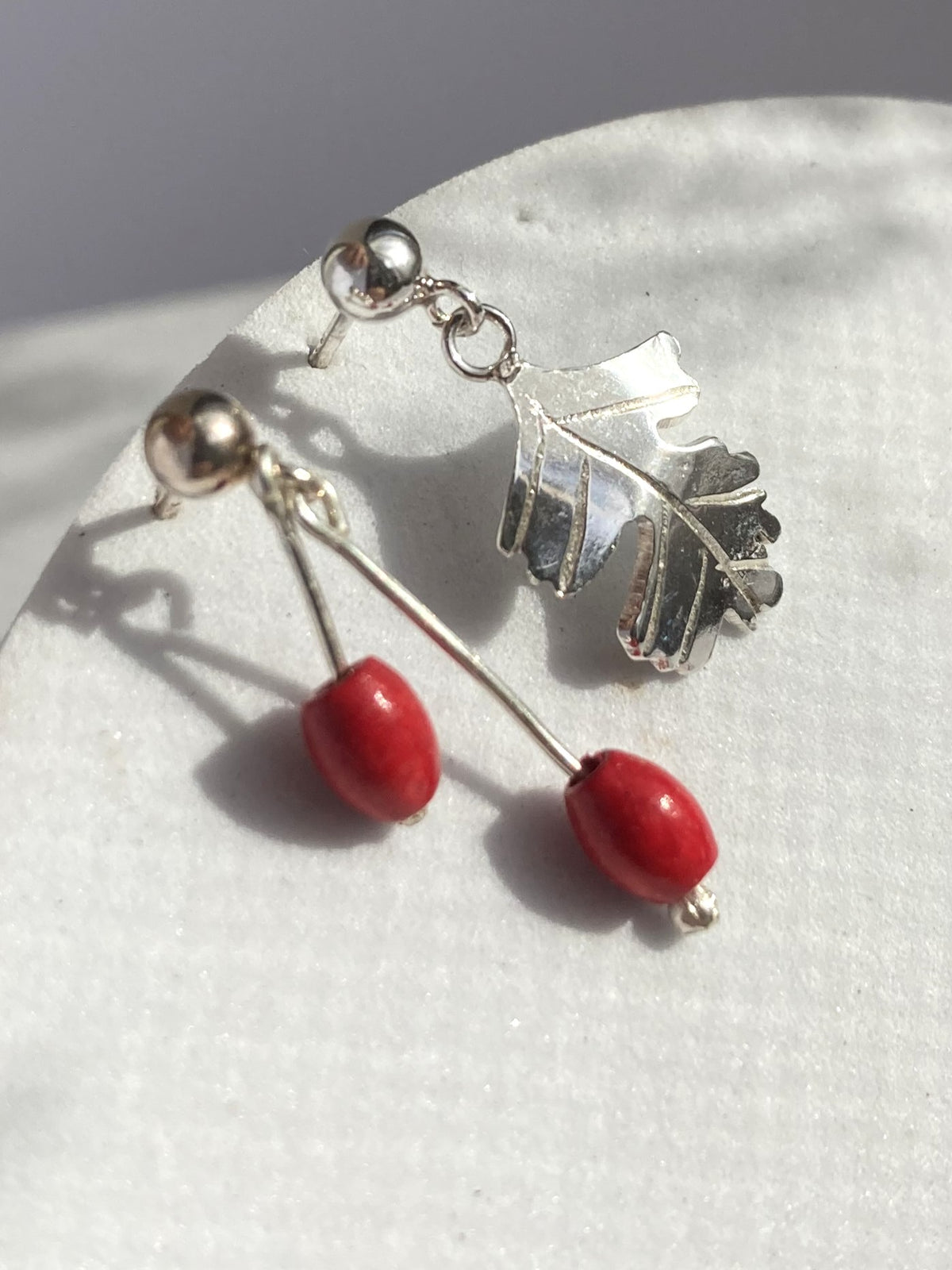A pair of handmade silver drop earrings with a Hawthorne leaf-shaped charm and red bead drops, displayed on a white textured surface.