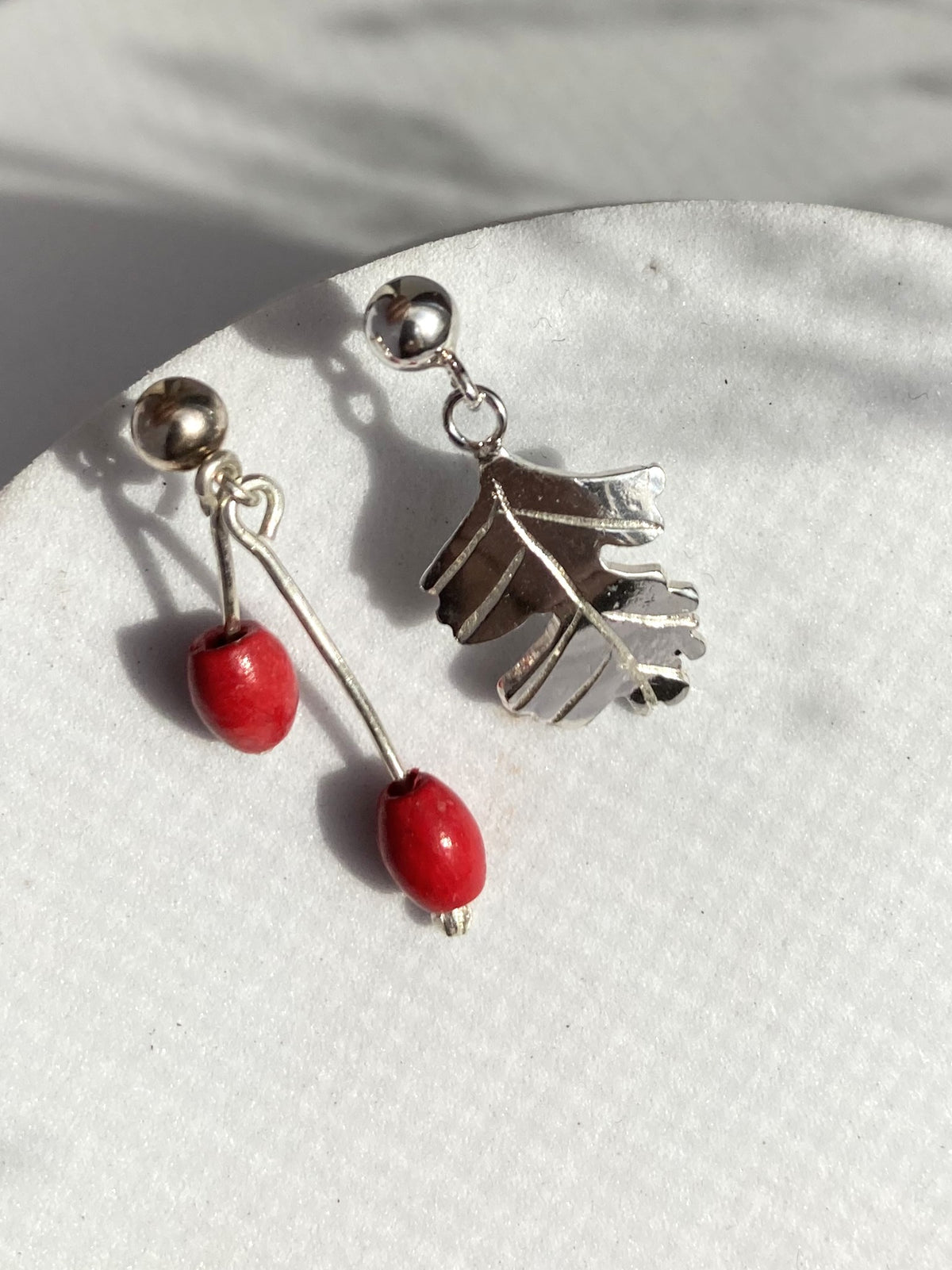 A pair of dangling earrings displayed on a white textured surface. One earring has a silver  Hawthorne leaf charm, and the other has a silver ball stud with a dangling silver wire ending in a red bead. By Kim Styles Jewellery