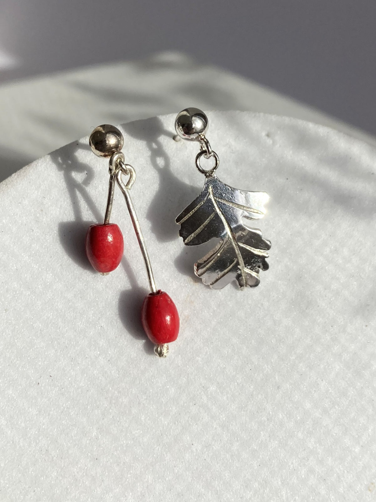 A pair of dangling earrings displayed on a textured white surface. One earring features a silver Hawthorne leaf charm, and the other features a silver post with two small red bead drops.