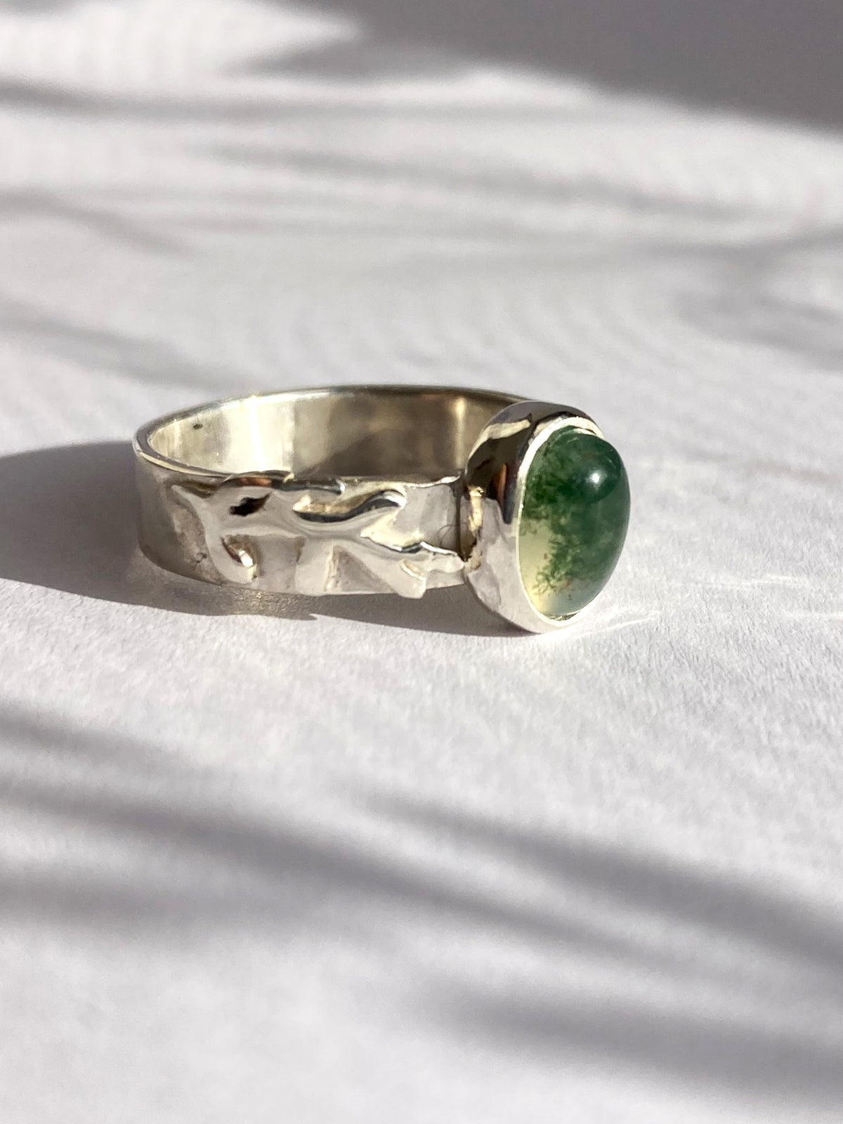 A silver ring with a textured, organic-shaped band and a round green gemstone, resting on a white textured surface with soft leaf shadows.